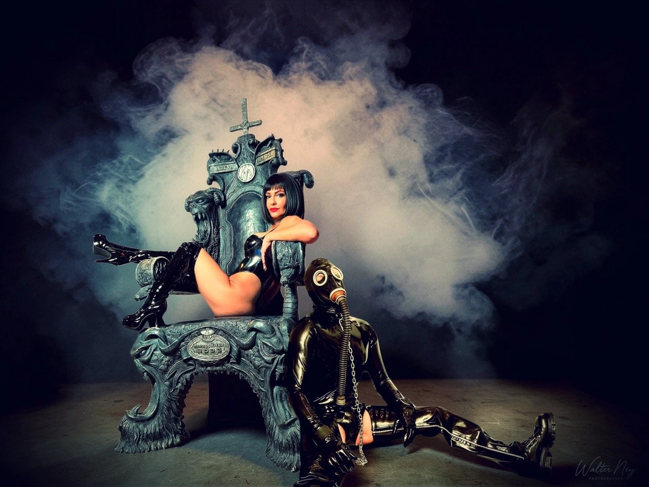 Domina auf dem Thron im Nebel, Sklave im Latexanzug mit Gasmaske zu ihren Füßen